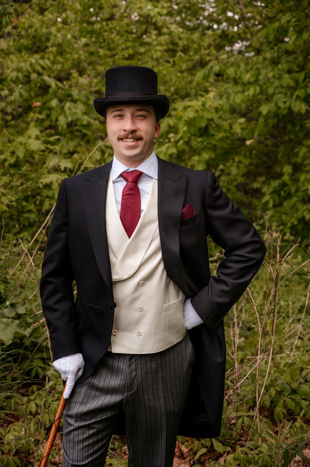 Dapper groom poses in top hat, morning coat, cream waistcoat, and burgundy tie among green foliage.