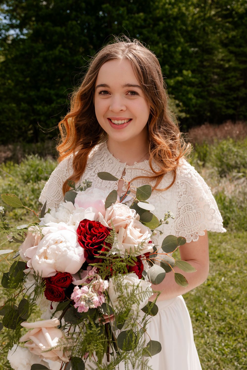 Radiant bride smiles holding cascading bouquet of white peonies and red roses in sunny outdoor setting.