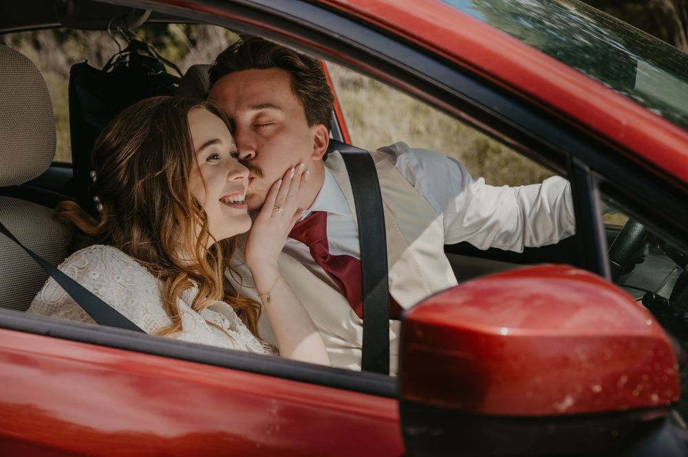 Bride and groom share a romantic kiss inside a red car on their wedding day.