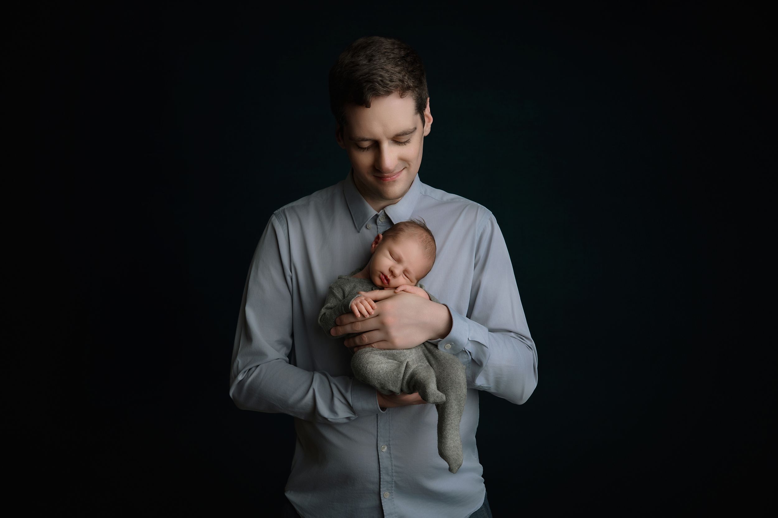Silhouetted against dark background, adult in gray sweater tenderly holds sleeping newborn baby.