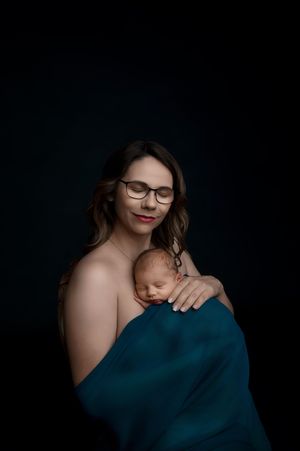 Intimate portrait of parent and sleeping newborn baby draped in deep teal fabric against dark background.