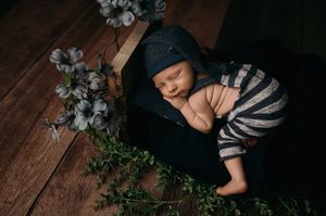 A sleeping infant lies peacefully on dark wooden flooring beside delicate white flowers and green foliage.