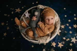 Baby wearing caramel colored outfit lies in round basket on dark backdrop with scattered gold stars.
