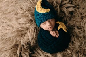 Sleeping baby wrapped in dark teal knit swaddle and matching hat with gold moon detail on fluffy fur backdrop.