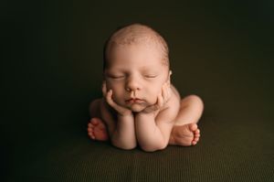 Sleeping newborn baby curled up in froggy pose on dark green textured backdrop with soft moody lighting.
