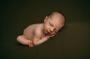 Sleeping newborn baby curled up peacefully on dark green textured blanket in professional studio portrait.