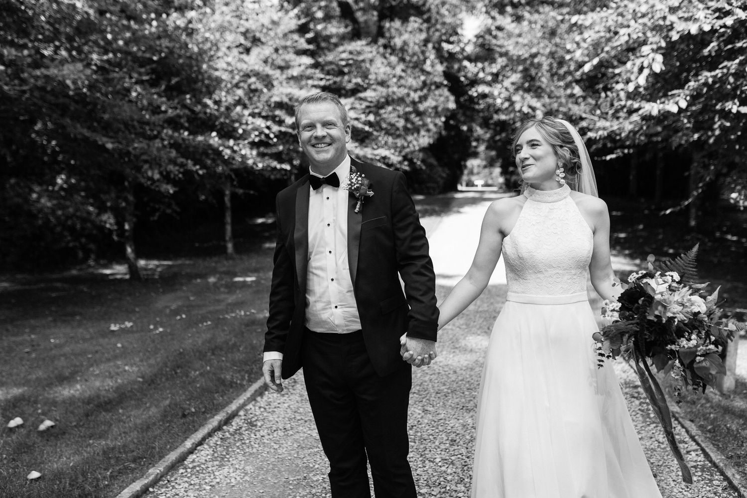 A couple walks hand in hand through a garden path on their wedding day wearing formal black tuxedo and white gown.