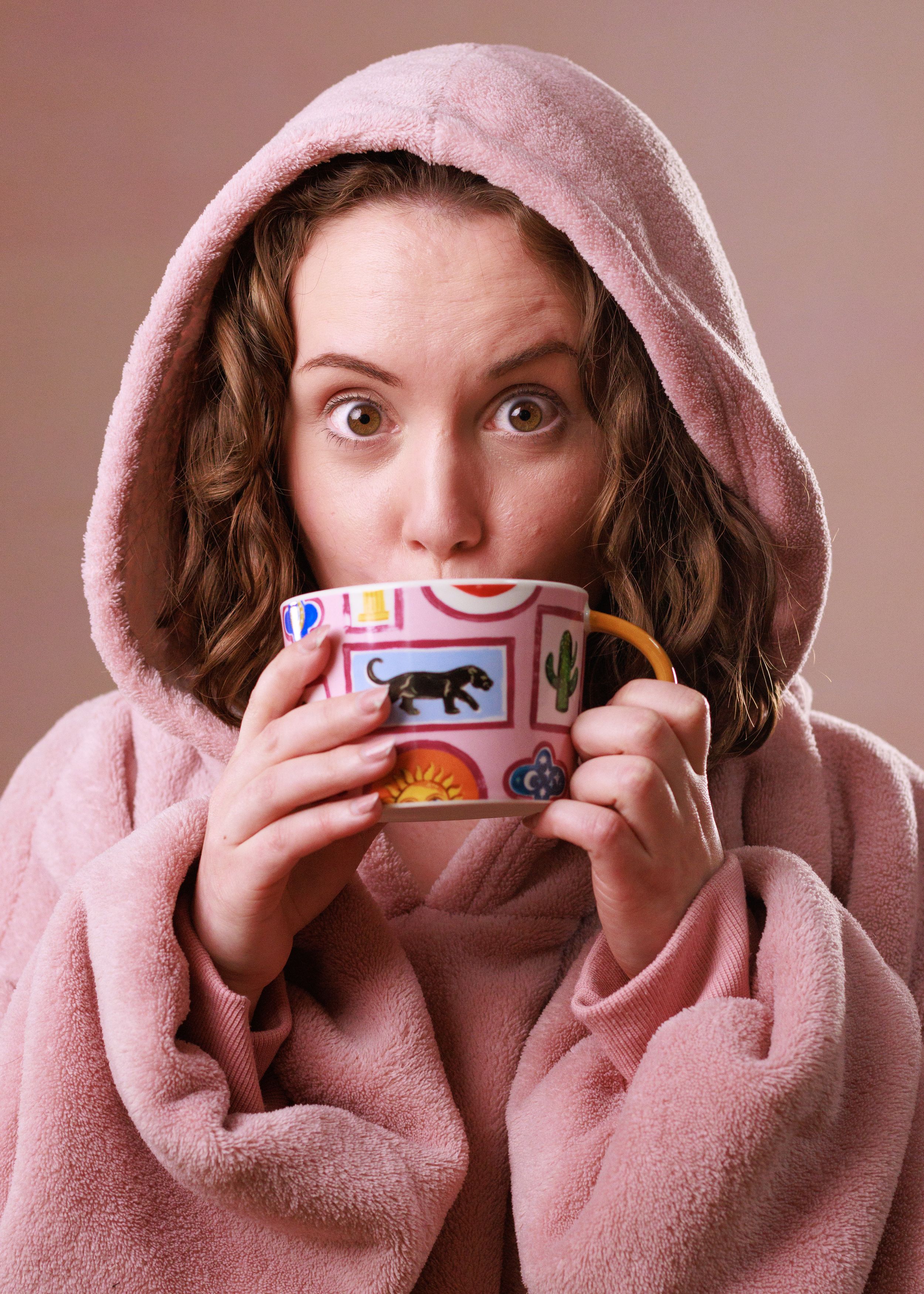 Someone wearing a cozy pink hooded bathrobe holds a decorative coffee mug with both hands while looking surprised.
