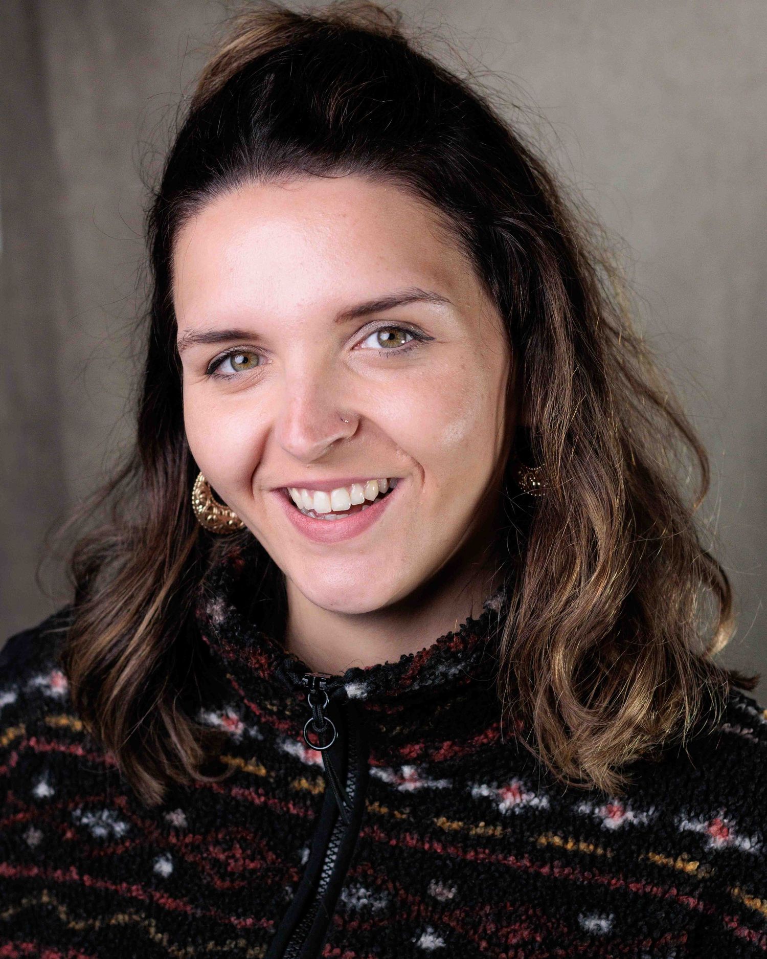 Someone with tousled brown hair wearing a black and white patterned winter sweater looks at the camera with a warm expression.