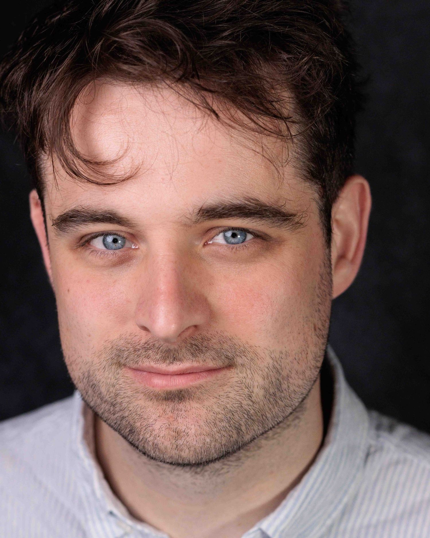 Professional headshot portrait of a person with bright blue eyes and dark tousled hair against a black background.
