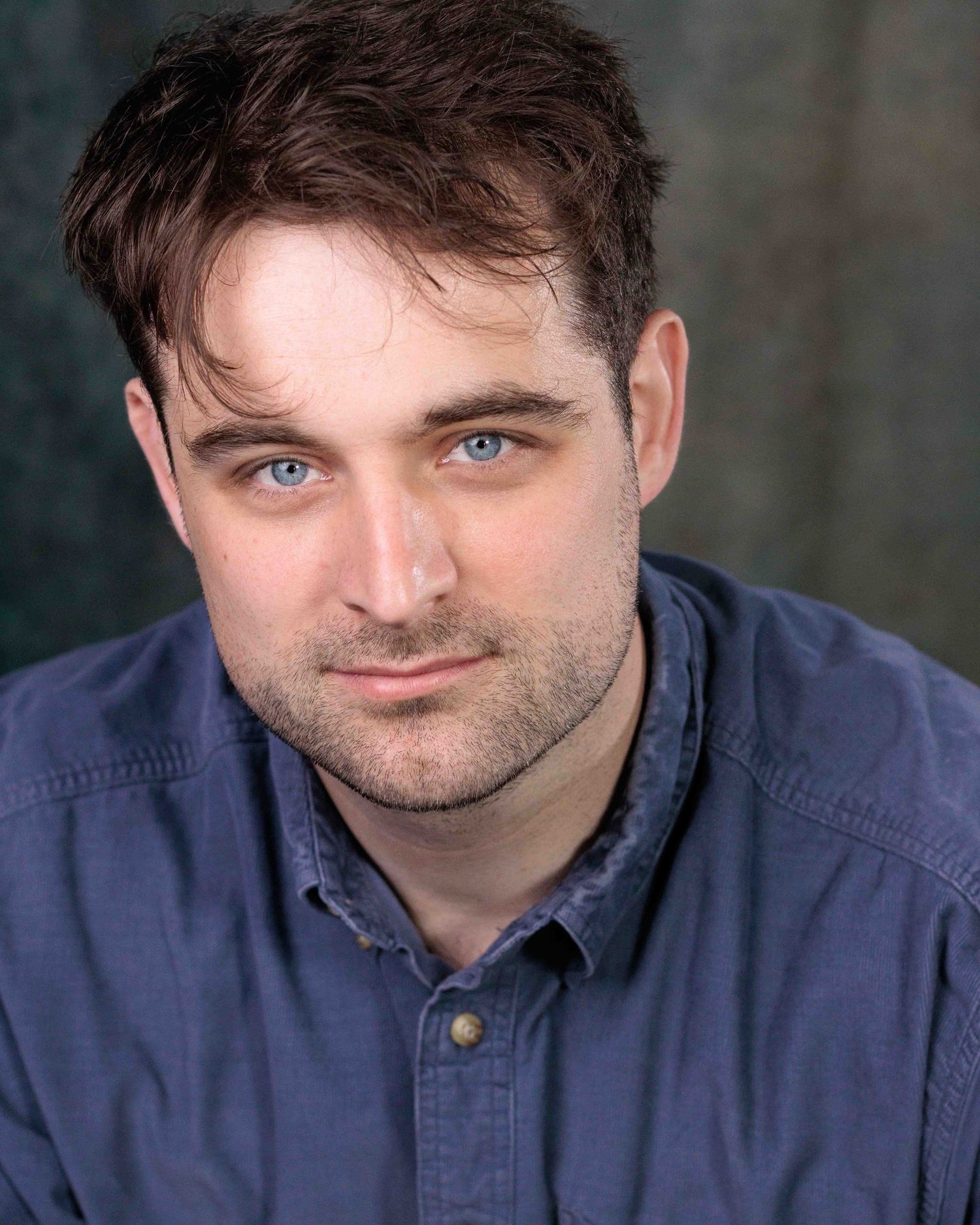 Portrait photo showing someone with bright blue eyes wearing a navy button-up shirt against a blurred dark background.