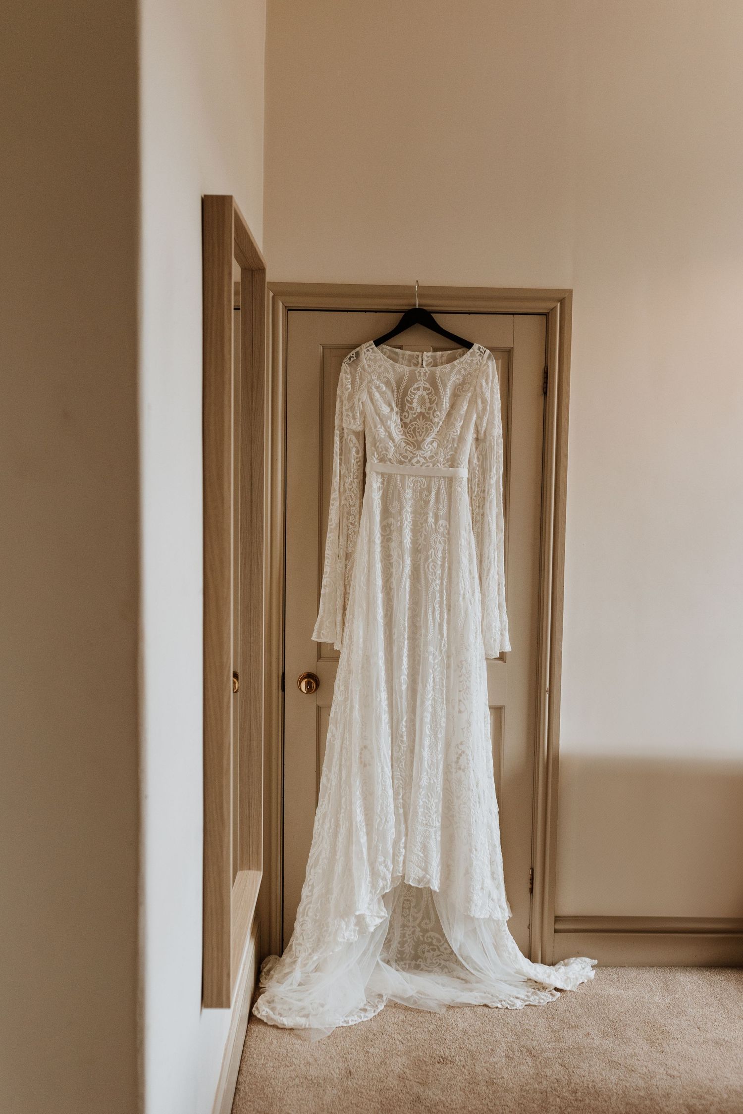 A delicate white lace wedding gown with long sleeves hangs elegantly against a neutral wall in soft natural light.