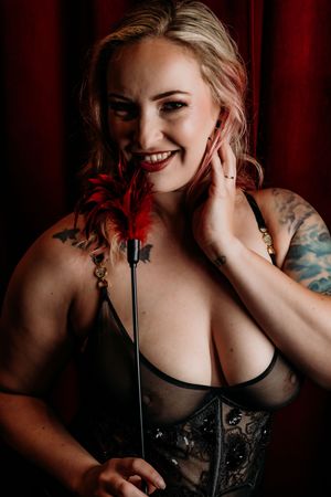Boudoir portrait with red feather prop against dramatic red curtain backdrop showcasing tattoo art. Grand Rapids Intimate  
Photography.