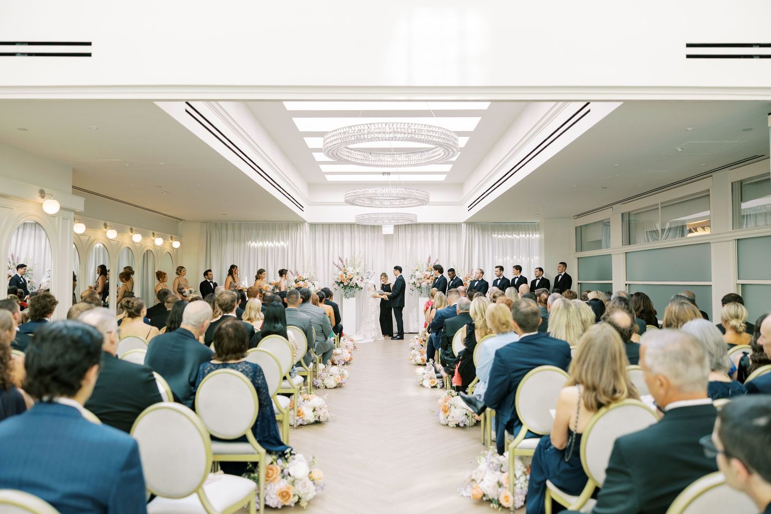 Wedding ceremony in a bright white ballroom with guests seated in rows and white floral decorations along the aisle.