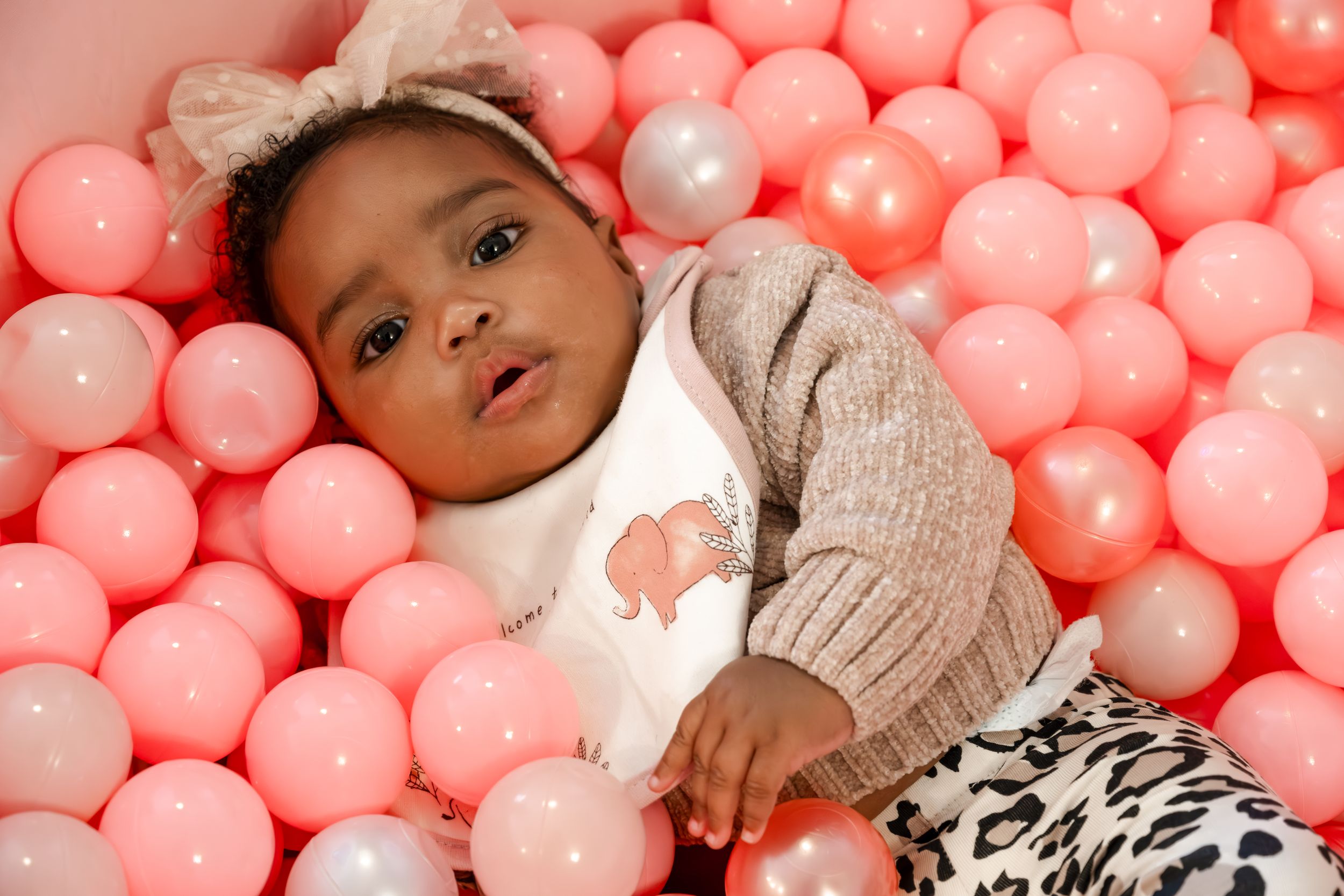 Baby in beige sweater lies in pink ball pit filled with coral colored and white plastic spheres.