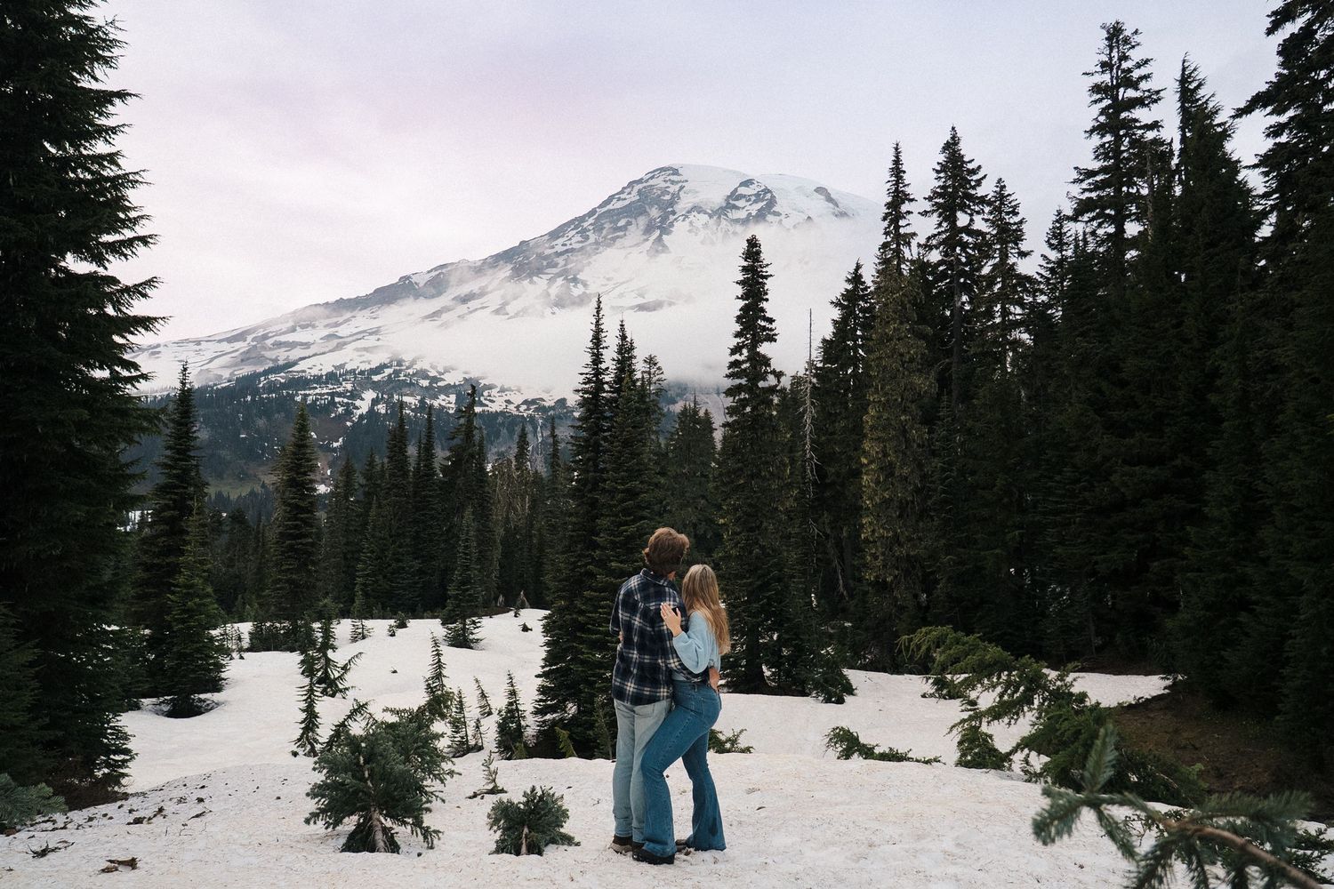 Two people stand together on a snowy mountain path with evergreen trees and snow-capped peaks in the background.