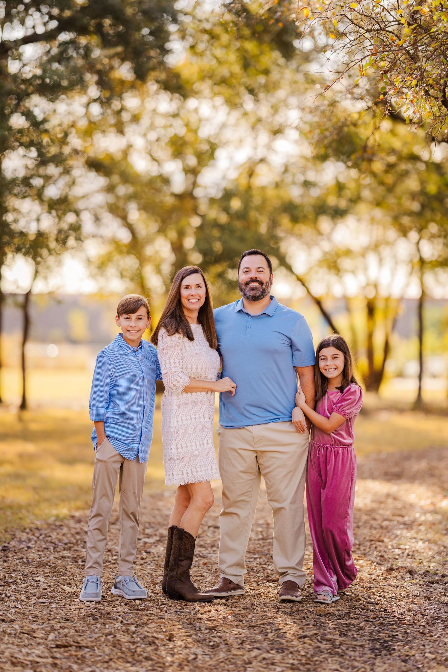 Family of four smiling together on a sunlit tree-lined path in warm golden light.