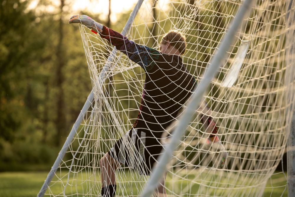 Youth soccer goalkeeper in white jersey leaps to block shot at net during sunset practice session.