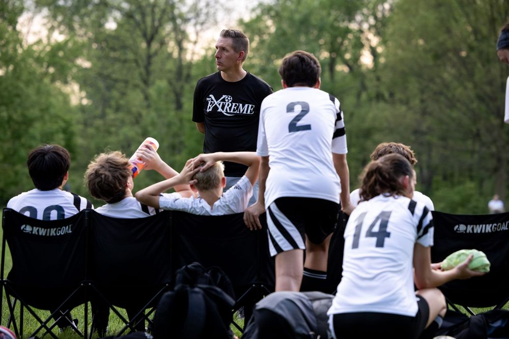 Athletes rest on sideline benches during outdoor soccer game while coach gives instructions at dusk.