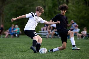Two youth soccer players battle for the ball during a match on a grass field with spectators in the background.