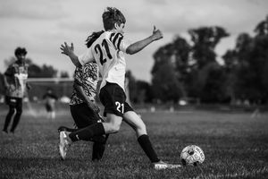 Black and white soccer action shot shows aggressive slide tackle between two players competing for the ball on a grass field.