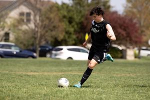 Soccer player in black uniform kicks the ball on a grass field during a match in autumn weather.