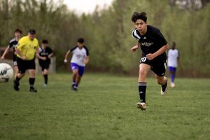 Soccer player in black uniform chases after ball during outdoor match on grass field with other players in background.