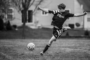 Black and white photo of a young soccer player kicking a ball on a grassy field while wearing a dark uniform.