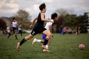 Two soccer players chase after the ball during an intense outdoor match on a grassy field.