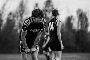 Soccer player in black uniform bends forward catching their breath during intense match at dusk.