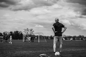 A person stands with hands on hips overlooking a soccer field during a youth game in black and white photography.