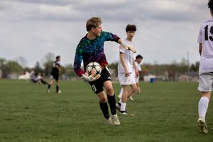 Soccer goalkeeper in a teal jersey jumps to make a save during a youth match on a grassy field.