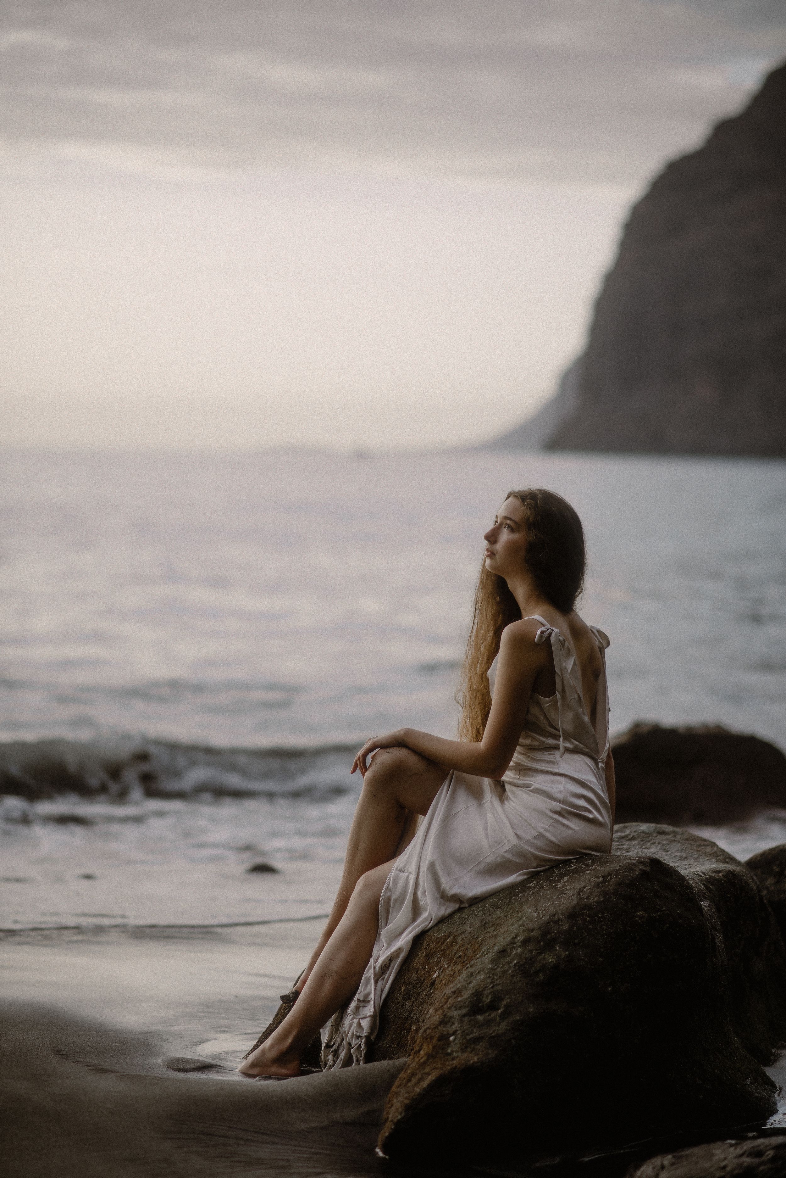 A person in a white dress sits peacefully on coastal rocks at sunset with cliffs and ocean waves in the background at Los Gigantes.