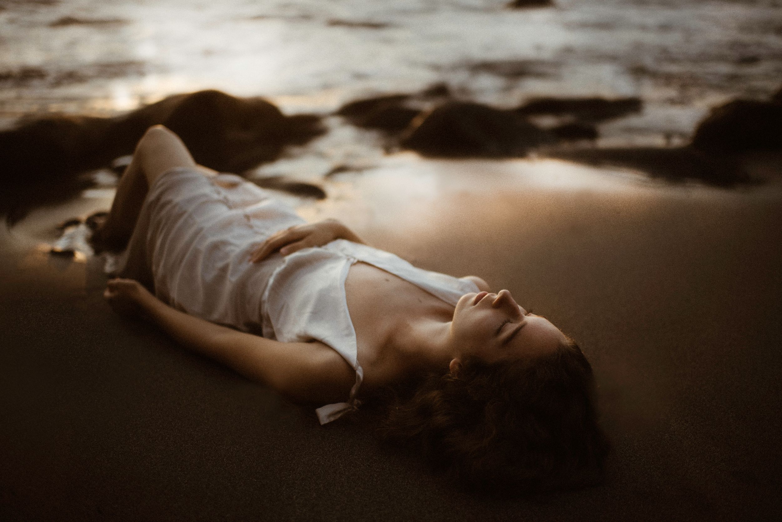 Artistic photograph of a person in a white satin dress lying on wet sand at sunset with moody, dramatic lighting at Los Gigantes, Tenerife.