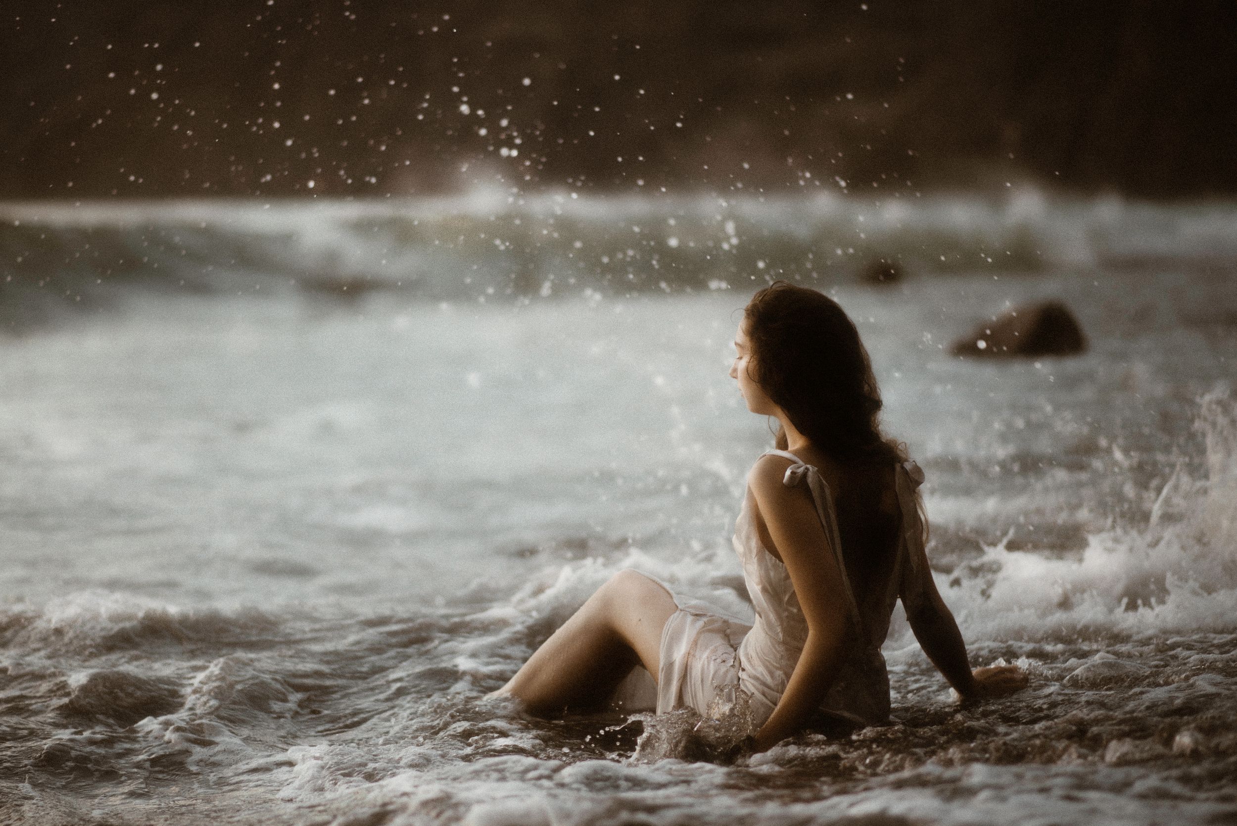 A moody seascape photo shows a figure sitting in splashing ocean waves at sunset wearing a light dress, at Los Gigantes, Tenerife.
