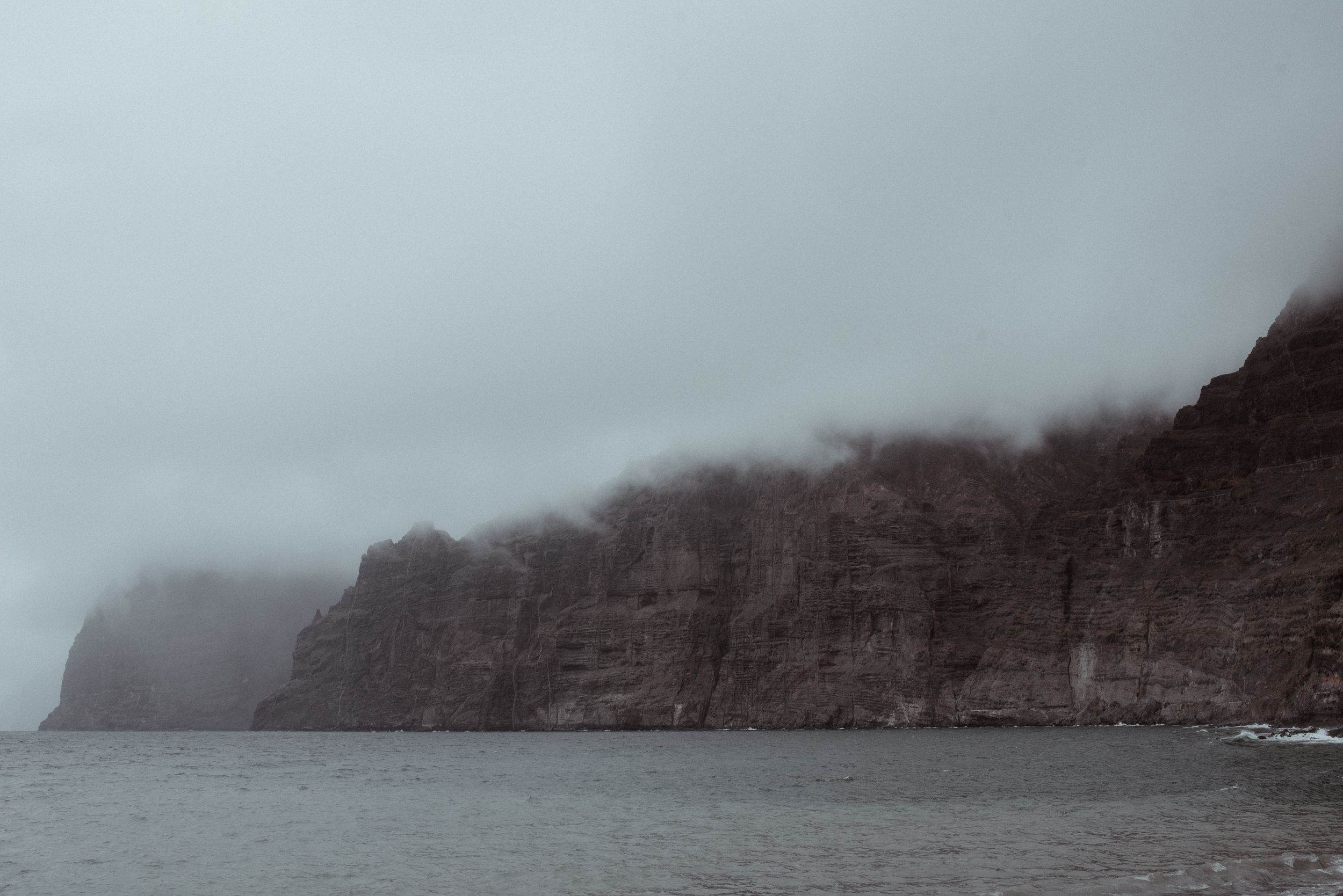 A dramatic coastline shrouded in fog with steep cliffs rising from the sea and misty atmosphere of Los Gigantes.