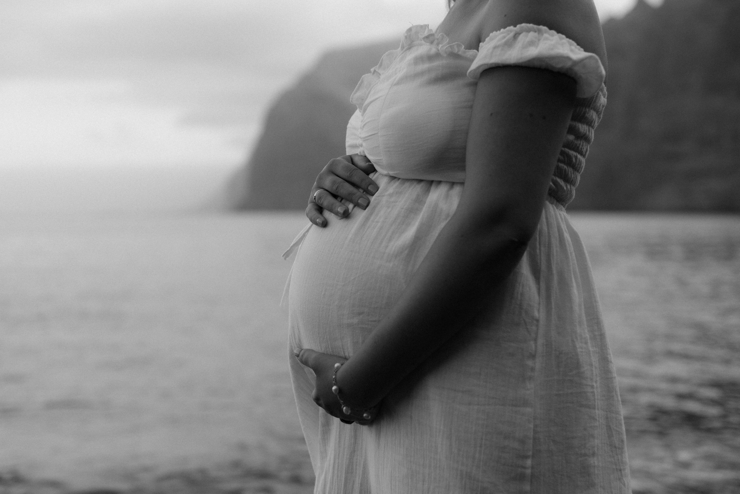 A pregnant person wearing a white flowing dress holds their belly while standing by a calm waterfront at Playa de los Guios, Tenerife.