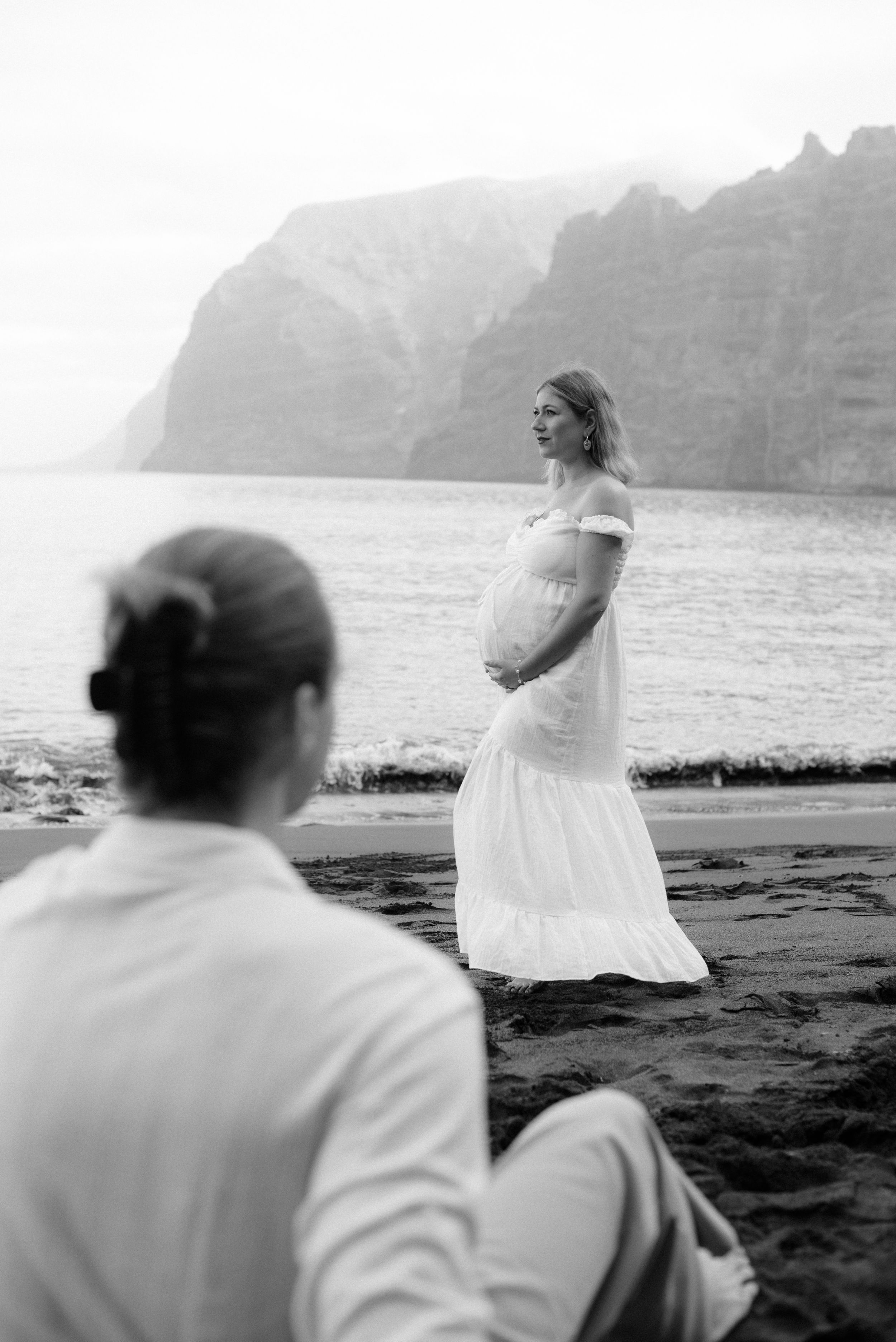 Black and white seaside maternity photo with dramatic cliffs in the background and a figure in a white dress on the beach of Los Gigantes.