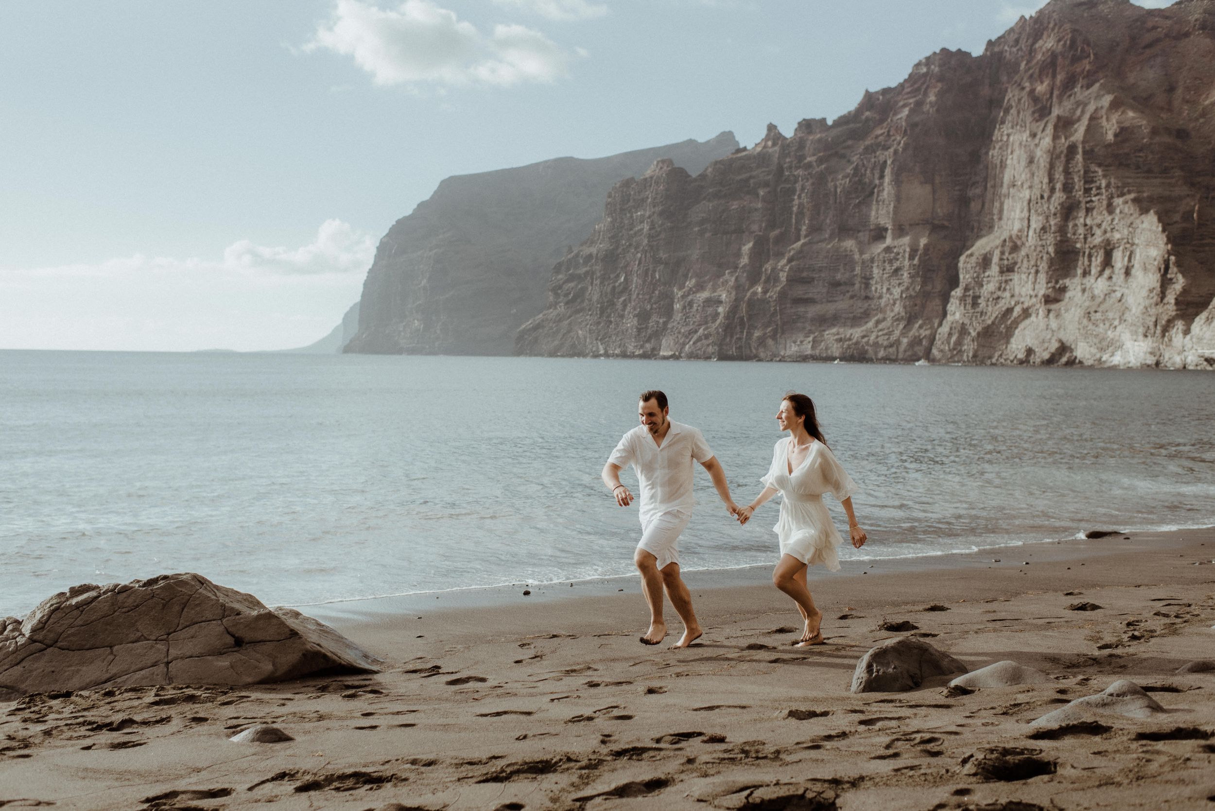 Two people in white outfits run together along a scenic beach with dramatic cliffs and ocean views of Los Gigantes.