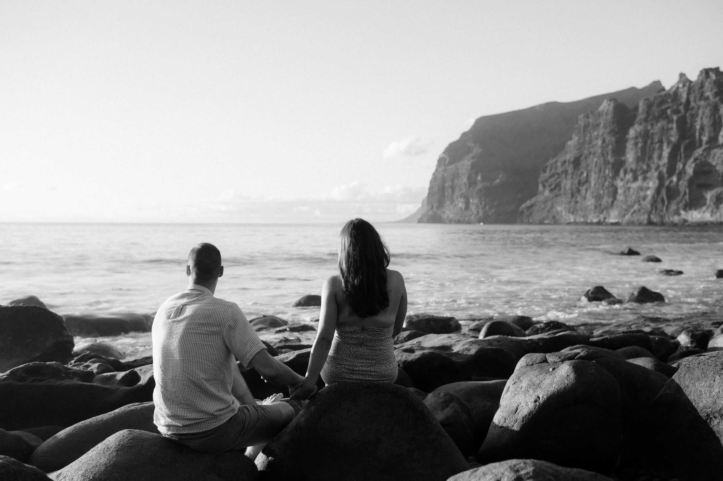A black and white photo of two people sitting on coastal rocks overlooking dramatic ocean cliffs.