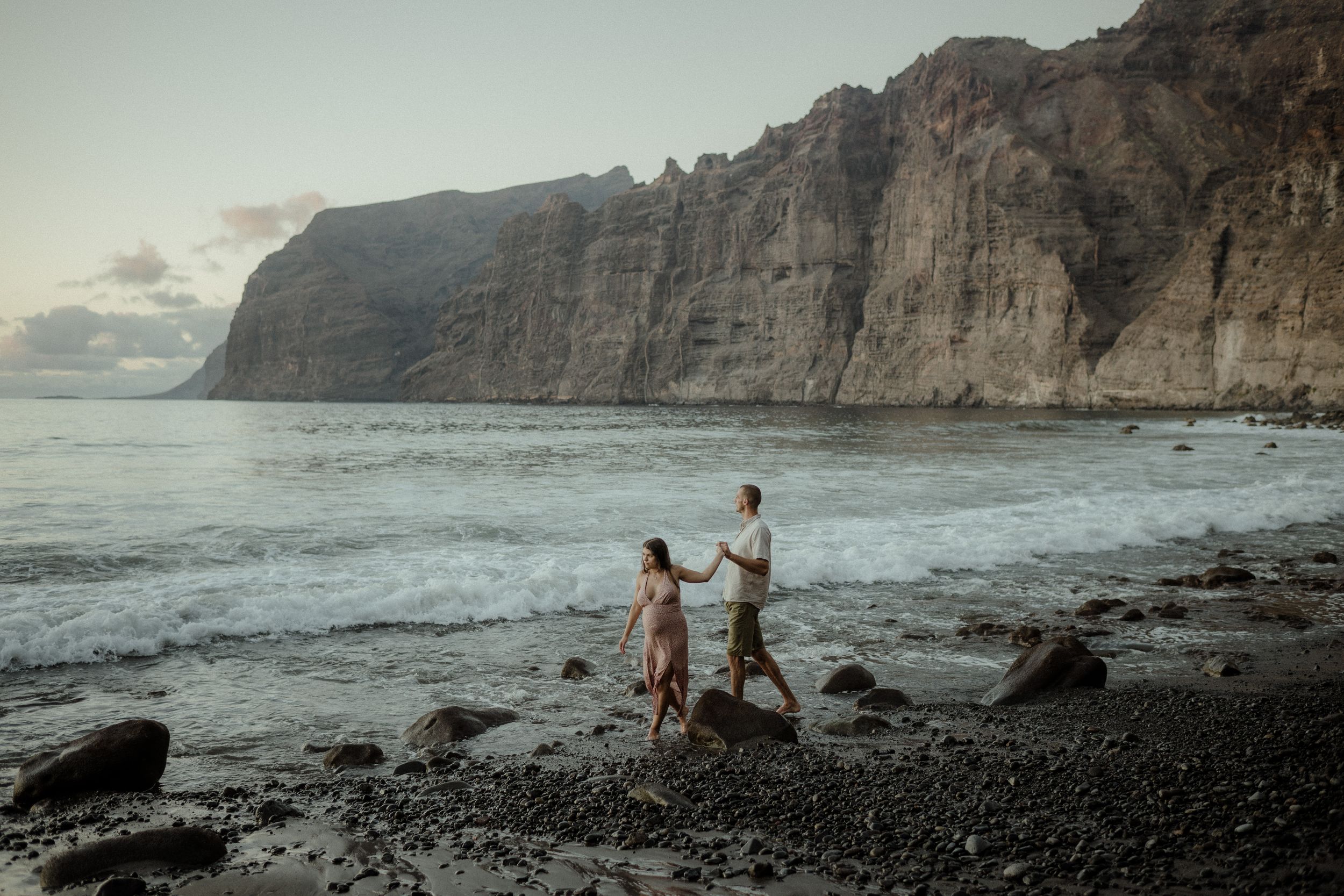 Couple walking together on rocky volcanic beach with towering Los Gigantes cliffs in background at dusk.