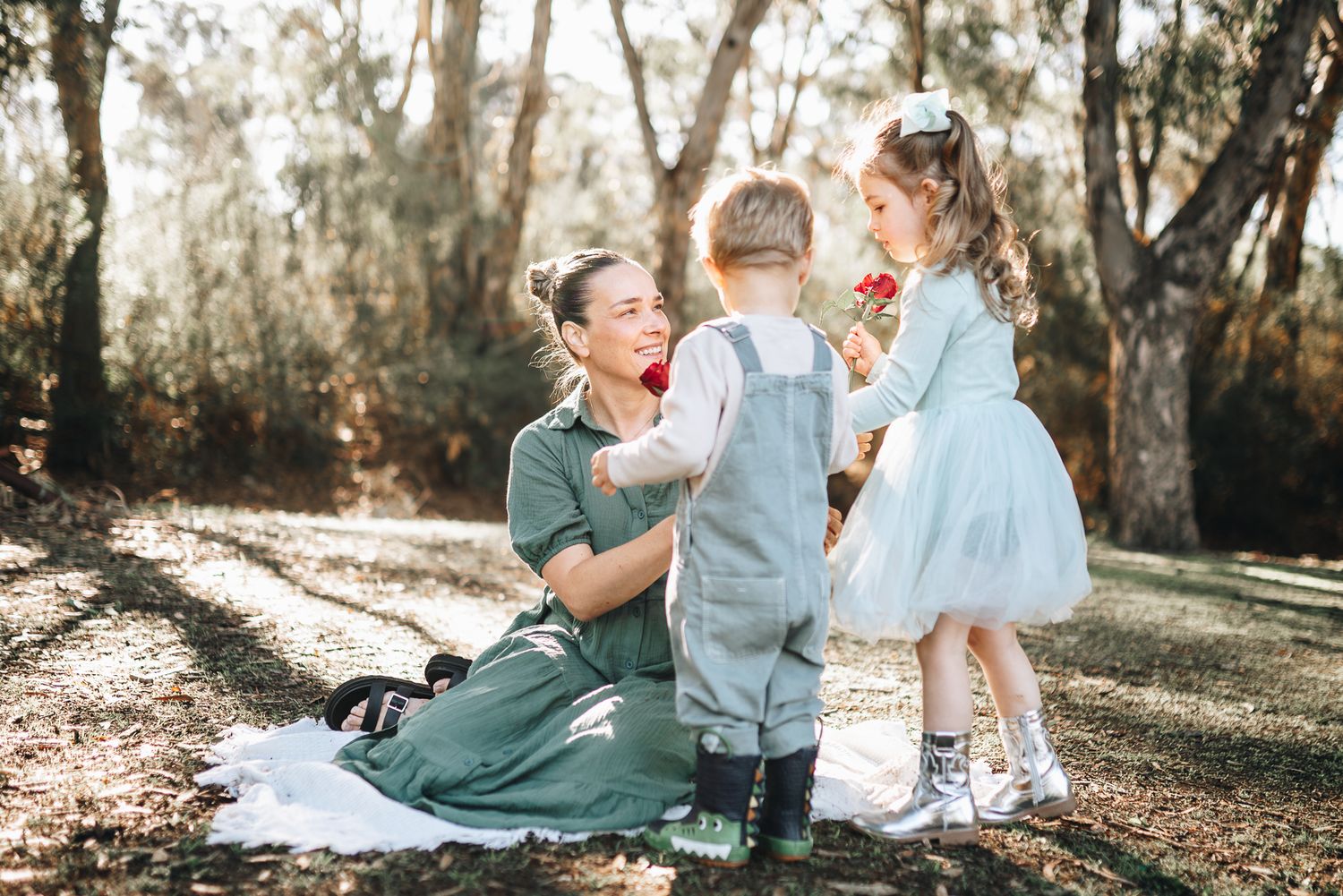 A mother in a teal dress and children in white clothing share tender moments together in a sunlit area by melbourne family photographer