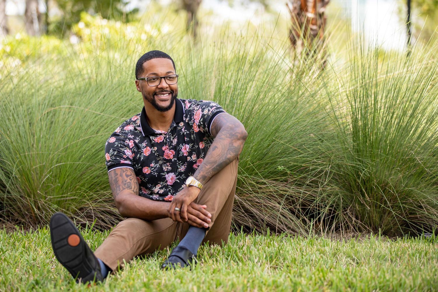 Person sitting on grass laughing in floral shirt and shorts.