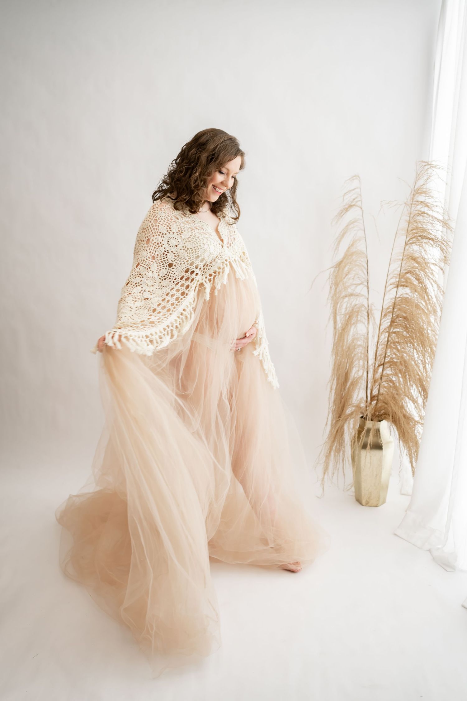 Beautiful maternity photo with flowing peach tulle gown and crochet shawl in bright minimalist studio setting with pampas grass.