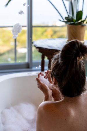 Woman relaxing in a bubble bath, blowing soap bubbles, surrounded by glass walls with an outdoor view from her Luxury Boutique Suite.