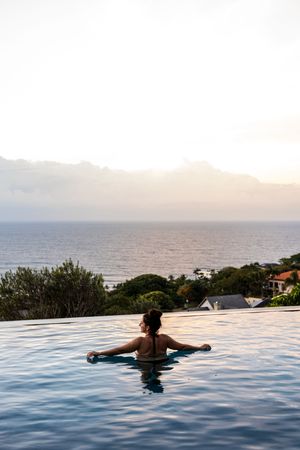 Woman relaxing in an infinity pool overlooking a serene ocean and mountain horizon at sunrise.