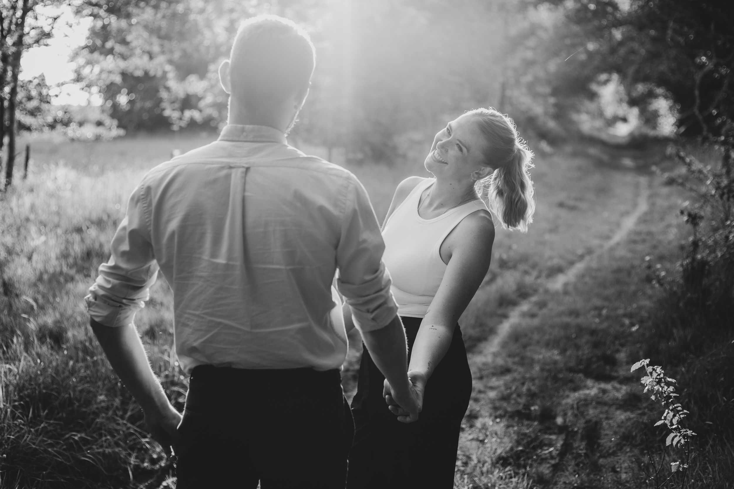 A couple walks hand in hand down a sunlit dirt path through nature in black and white.