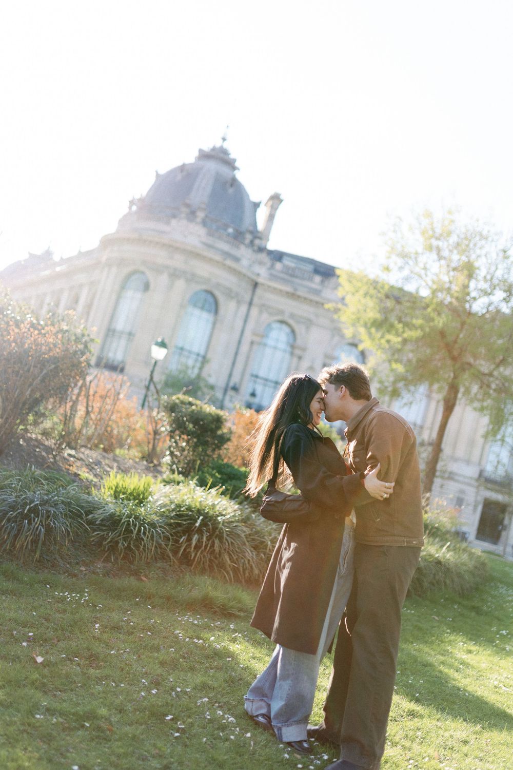 A romantic couple share an intimate moment in front of an ornate historic building with a domed roof on a sunny afternoon.