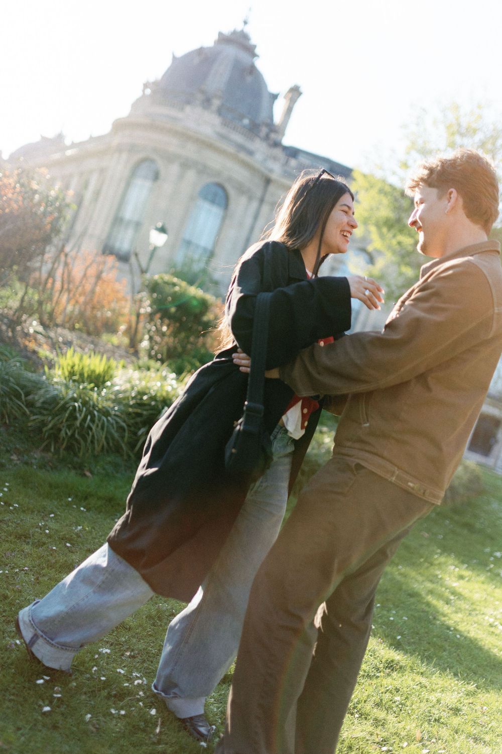 A couple laughs together while embracing on a sunny day outside a grand historic building with ornate architecture.