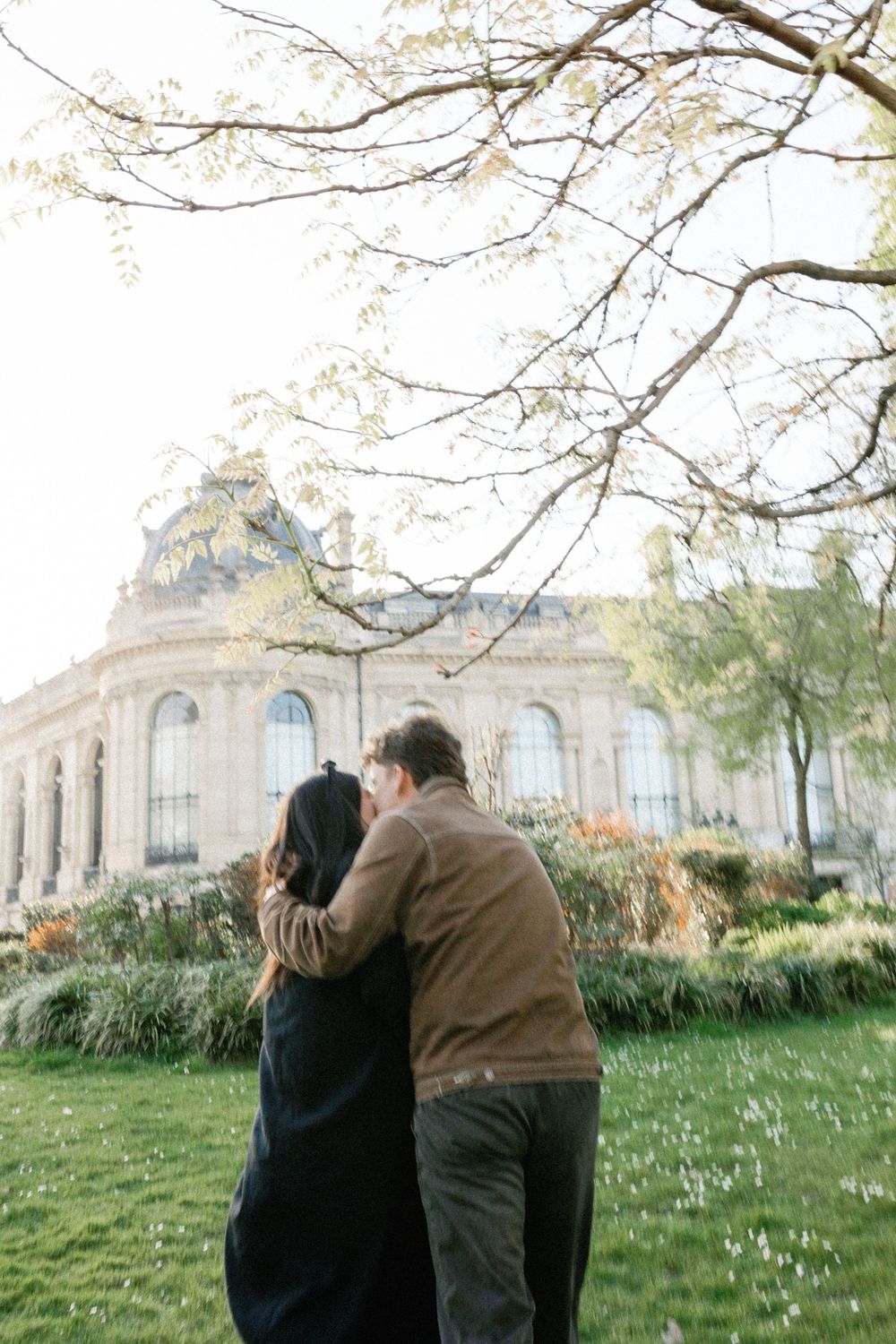 A couple embraces on a grassy lawn in front of an elegant classical building with arched windows on an autumn day.